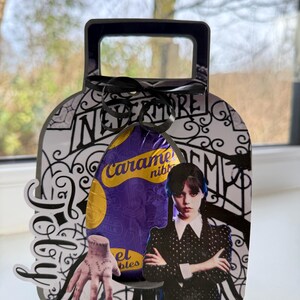 May include: A decorative box with a handle, in black and white, with the word "Nevermore" and a cartoon image of a character in a black dress. A purple candy bar is inside the box, and a black ribbon is tied at the top.