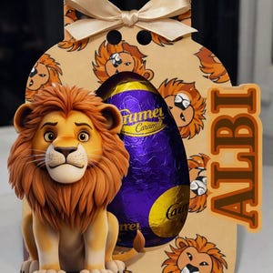 May include: A gift box with a cartoon lion and a purple foil-wrapped egg. The box is decorated with lion illustrations and a beige bow. The word "ALBI" is printed on the side of the box.