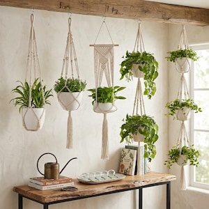 May include: Several macrame plant hangers with white pots holding green plants, suspended from a wooden beam. A wooden table displays books, a watering can, and a decorative tray. The room has a neutral colour scheme.