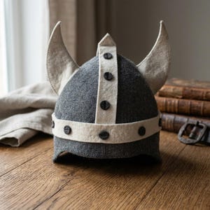 Könnte beinhalten: Ein grauer Wikingerhelm aus Filz mit cremefarbenen Akzenten und schwarzen Knöpfen. Der Helm hat zwei große, gebogene Hörner und ein cremefarbenes Band um die Basis. Der Helm wird auf einer Holzoberfläche präsentiert.