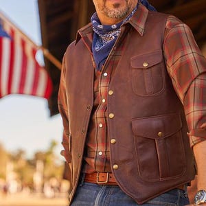 May include: A brown leather vest with a button-up front and two large pockets. The vest has a flap over the top pocket and a smaller pocket below. The vest is worn over a plaid shirt.
