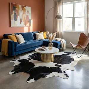 May include: A modern living room featuring a blue velvet sectional sofa with yellow and cream throw pillows and a matching throw blanket. A round coffee table with a marble top and gold base sits on a black and white cowhide rug. A brown leather butterfly chair is in the corner.