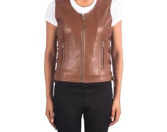 May include: A brown leather vest with a zip-up front and two zippered pockets. The vest has decorative lacing on the sides. The model is wearing a white t-shirt and black pants.