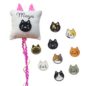 Peut inclure: Un oreiller blanc avec des oreilles roses et le nom "Maya" brodé dessus. Un visage de chat noir est brodé sur l'oreiller. L'oreiller est attaché à une ficelle bouclée rose. Il y a neuf visages de chats brodés de différentes couleurs et motifs autour de l'oreiller.
