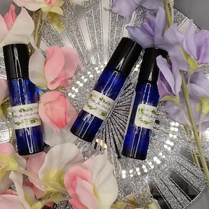 May include: Three blue glass roller bottles with black caps. Each bottle has a white label with the text "Setting the Mood Oil" in black lettering. The bottles are on a silver background with pink and purple flowers.