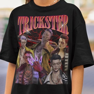 Könnte beinhalten: Schwarzes T-Shirt mit dem Wort "TRICKSTER" in einer Retro-Schriftart über einer Grafik von mehreren Charakteren. Die Grafik hat einen Vintage-Look mit rotem Blitzhintergrund.