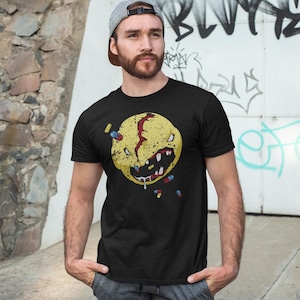 Puede incluir: Camiseta negra con un diseño de cara sonriente amarilla distorsionada, con una cicatriz roja y dientes afilados. Las pastillas salen de la boca y rodean la cara. La camiseta es de cuello redondo.