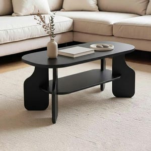 May include: A black, modern coffee table with an oval top and a lower shelf. The table has a minimalist design and is made of wood. A vase with dried flowers, a book, and a small plate are on the table. The table is 40 inches long and 20 inches wide.