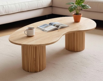 May include: A light-colored wooden coffee table with a unique kidney bean shape. The table has two cylindrical legs made of vertical wooden dowels. A mug and an open book rest on the table's surface, and a small potted plant sits nearby.