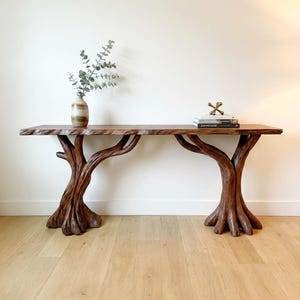 May include: A long wooden console table with a natural live edge top supported by two base structures shaped like tree trunks with visible roots. A ceramic vase with eucalyptus branches and a stack of books with a gold jack object sit on top.