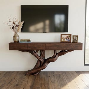 May include: A dark brown wooden console table featuring a flat rectangular top with three drawers and a base sculpted to resemble the trunk and spreading branches of a tree. The table holds a ceramic vase with dried botanicals and two frames.