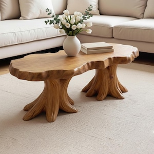 May include: A wooden coffee table with a natural edge design, supported by two tree trunk-shaped bases. The table is light brown and has a smooth surface. A white vase with white flowers and a stack of books are on top.