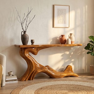 May include: A handcrafted wooden console table with a unique, organic design. The table features a natural wood finish and a sculptural base. Decorative vases and a framed artwork are displayed on the table's surface. A round jute rug is on the floor.