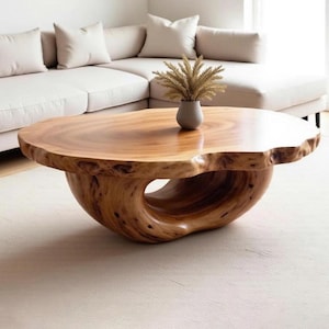 May include: A unique, solid wood coffee table with a natural edge. The table has a smooth, polished surface and a distinctive, sculptural base with a large, open circular design. A small vase with dried plants sits on top.