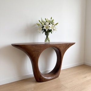 Può includere: Un tavolo console in legno marrone scuro con una silhouette liscia e curva caratterizzata da un ampio intaglio circolare al centro. Un vaso in vetro trasparente contenente un bouquet di gigli bianchi e fogliame verde poggia sulla superficie.