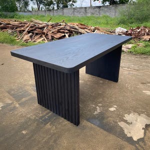 May include: A modern, rectangular black wooden table. The tabletop has rounded corners and a smooth surface. The table is supported by a base with vertical, ribbed detailing on one side and a solid panel on the other. The table is suitable for various uses.
