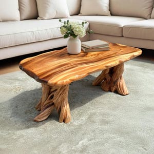 May include: A handcrafted wooden coffee table with a natural edge. The table features a warm, brown finish and is supported by intricately carved tree trunk legs. A white vase with flowers and a stack of books rest on the table's surface.