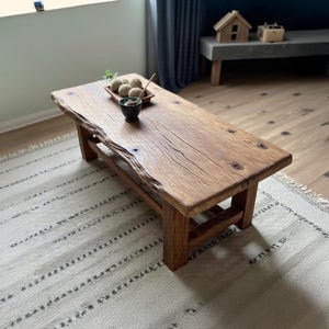 Puede incluir: Una mesa de centro rectangular de madera rústica con borde natural. La mesa es de madera marrón claro y tiene una veta visible. Encima hay una pequeña bandeja de madera con objetos decorativos. La mesa mide 127 cm de largo.