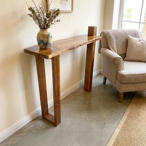 May include: A tall, narrow wooden console table with a natural live edge top and a smooth, polished brown finish. The table features two sturdy, rectangular legs and supports a ceramic vase filled with dried branches and foliage.