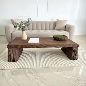 Könnte beinhalten: Ein rechteckiger, dunkelbrauner Couchtisch aus Holz mit strukturierter Oberfläche und Beinen im Baumstamm-Stil. Der Tisch steht vor einem beigen Sofa mit passenden Kissen. Eine kleine Vase mit Grün und eine dekorative Schale stehen auf dem Tisch.