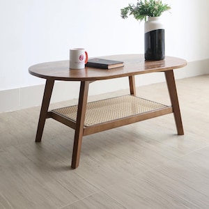 Puede incluir: Mesa de centro ovalada de madera con un estante inferior con un diseño de ratán tejido. La mesa tiene cuatro patas anguladas y está coronada con un libro, una taza y un jarrón con vegetación. La mesa es de color marrón cálido.