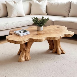 May include: A distinctive coffee table crafted from natural wood, featuring an organic, freeform design. The table has a light brown finish and is supported by three tree trunk-style legs. A small plant and a book are placed on the surface.