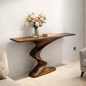 Op de afbeelding: Een donkerbruine, sculpturale console met een unieke, golvende steun. De tafel bevat een glazen vaas met roze en witte bloemen en een klein boek. De tafel is ongeveer 152 cm lang.