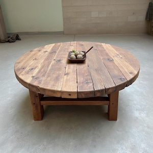 Puede incluir: Una mesa de centro redonda y rústica hecha de madera reciclada. La mesa tiene un acabado de madera natural con nudos y vetas visibles. Se apoya sobre cuatro patas de madera robustas. Una pequeña bandeja de madera con esferas decorativas descansa sobre la mesa.
