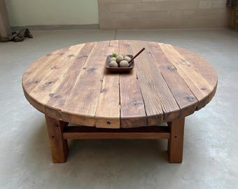 Può includere: Un tavolino da caffè rotondo e rustico in legno di recupero. Il tavolo ha una finitura in legno naturale con nodi e venature visibili. Poggia su quattro robuste gambe in legno. Un piccolo vassoio di legno con sfere decorative è posto sulla superficie del tavolo.