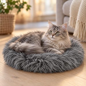Puede incluir: Una cama redonda y esponjosa para mascotas de color gris con una textura gruesa de piel sintética. Un gato atigrado de pelo largo con pelaje gris y marrón está acurrucado en el centro de la cama, descansando sobre el material suave y afelpado.