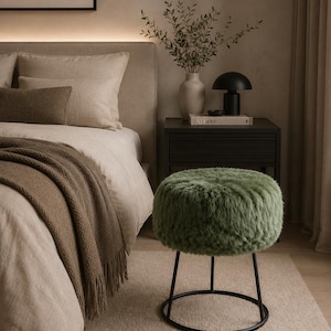 Pode incluir: Um banco redondo com uma almofada de assento em pele sintética verde-sálvia, apoiado por uma estrutura de metal preto com base circular. O banco está posicionado num quarto ao lado de uma cama com roupa de cama neutra e uma mesa de cabeceira.