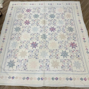 Puede incluir: Una colcha de retazos blanca y crema con un patrón de estrellas. La colcha tiene un borde azul y rosa con un patrón de diamantes.