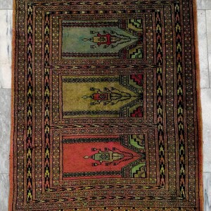 Könnte beinhalten: Rechteckiger Teppich mit drei zentralen Feldern in Grün, Rot und Gold. Der Teppich hat einen detaillierten Rand mit geometrischen Mustern in Schwarz, Braun und Grün. Der Teppich ist wahrscheinlich aus Wolle.