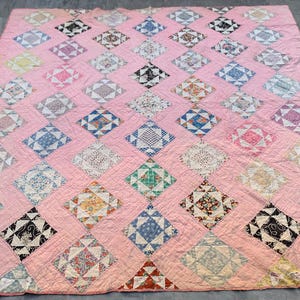 May include: A pink quilt with a diamond pattern. Each diamond is made of a different fabric with various colors and patterns. The quilt is made of many different fabrics, including floral prints, geometric patterns, and solid colors.