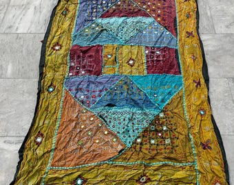Op de afbeelding: Een kleurrijk patchwork-deken met een geometrisch ontwerp. Het deken is gemaakt van verschillende stoffen in tinten blauw, groen, rood en bruin. Het deken is versierd met kleine spiegels en borduurwerk.