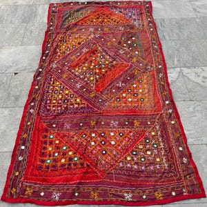 Könnte beinhalten: Eine rote, kastanienbraune und goldene Patchwork-Decke mit aufwendigen Stickereien und kleinen Spiegelakzenten. Die Decke ist aus einem weichen, strukturierten Stoff gefertigt und hat einen Fransenrand.