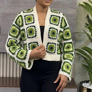 Könnte beinhalten: Eine cremefarbene Häkeljacke mit grünen quadratischen Mustern. Die offene Jacke hat einen V-Ausschnitt und lange Ärmel. Das Häkeldesign integriert Grün- und Cremetöne und erzeugt eine optisch ansprechende Textur. Dieses handgefertigte Kleidungsstück ist eine stilvolle Ergänzung für jede Garderobe.