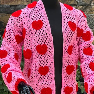 Könnte beinhalten: Eine rosa gehäkelte Strickjacke mit roten Filzherzen. Die Strickjacke hat eine lockere Passform und lange Ärmel.