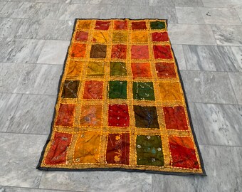 Op de afbeelding: Een rechthoekig patchworktextiel met een rasterpatroon van kleurrijke vierkanten. De vierkanten zijn in tinten rood, oranje, geel, groen en bruin, met gouden rand. Het textiel heeft een donkergrijze rand.