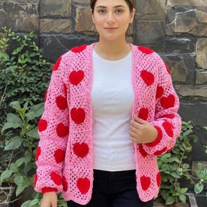 Könnte beinhalten: Ein rosa gehäkelter Cardigan mit großen roten Herzapplikationen. Der offen geschnittene Pullover hat lange Ärmel und wird über einem weißen Oberteil getragen. Das ist ein handgefertigtes, gemütliches Kleidungsstück.