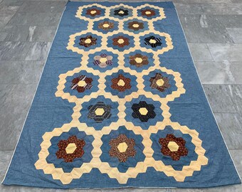Op de afbeelding: Een grote, handgemaakte quilt met een blauwe achtergrond en een herhalend bloemenpatroon. De bloemen zijn gemaakt van verschillende stoffen met patronen in tinten bruin, geel en zwart, geplaatst in zeshoekige vormen. De quilt is waarschijnlijk voor een bed of een groot oppervlak.