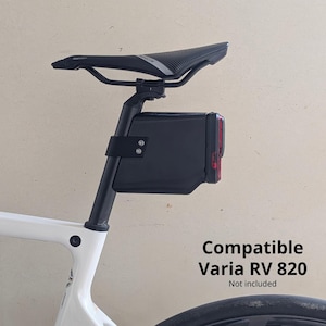 Peut inclure: Accessoire de vélo noir monté sur un tube de selle blanc. L'accessoire est de forme rectangulaire avec une lumière rouge sur le côté. Le texte "Compatible Varia RV 820" est visible.