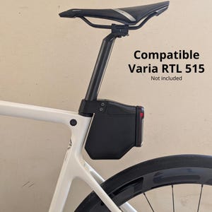Peut inclure: Une sacoche de selle de vélo noire montée sur la tige de selle d'un cadre de vélo blanc. La sacoche comporte un feu rouge orienté vers l'arrière. Le texte indique : Compatible Varia RTL 515. Not included. Le vélo a une selle et une roue noires.