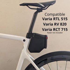 以下が含まれることがあります： 白い自転車フレームに取り付けられた黒いシートポストマウント。Compatible Varia RTL 515、Varia RV 820、Varia RCT 715、Radar not includedというテキストが表示されています。マウントは黒い自転車用サドルの下に配置されています。