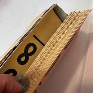 May include: A stack of rectangular cards with yellow edges and a tan cardboard holder. The cards feature a large black infinity symbol printed on the yellow surface. The edges of the cards are visible, showing a thin, layered paper construction.