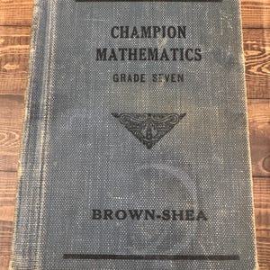 May include: A vintage textbook titled "Champion Mathematics Grade Seven" with a blue cover and black text. The book's cover has a textured pattern and the publisher's name, Brown-Shea, is printed at the bottom.