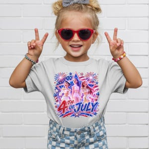 Puede incluir: Una ni&ntilde;a peque&ntilde;a lleva una camiseta gris claro con un dise&ntilde;o gr&aacute;fico colorido. El gr&aacute;fico presenta a tres mujeres con trajes blancos, fuegos artificiales y el texto "4th of July" en rojo y azul. La ni&ntilde;a lleva gafas de sol rojas.