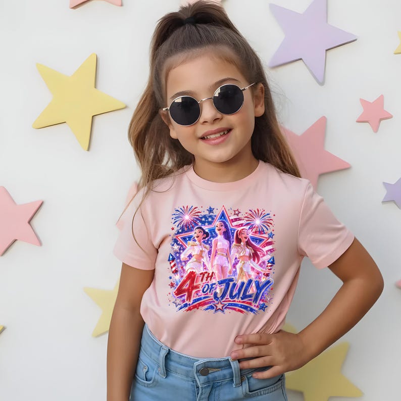 Puede incluir: Una persona joven que lleva una camiseta rosa claro con un dise&ntilde;o gr&aacute;fico que celebra el 4 de julio. El dise&ntilde;o presenta tres figuras, fuegos artificiales y el texto "4th of July". La persona tambi&eacute;n lleva gafas de sol redondas.