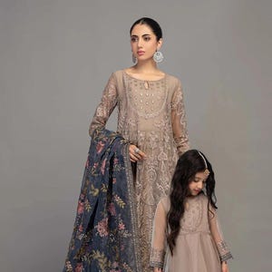 May include: Two women wearing matching beige embroidered dresses with long sleeves. The woman in the front is holding a blue floral embroidered shawl. The woman in the back is wearing a matching shawl with a floral design.