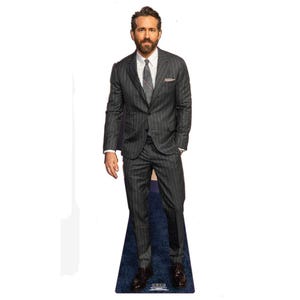 May include: A life-size cardboard cutout of a man in a dark gray pinstripe suit, white shirt, and gray tie. The cutout has a blue base and brown dress shoes. The man has a beard and is looking forward.